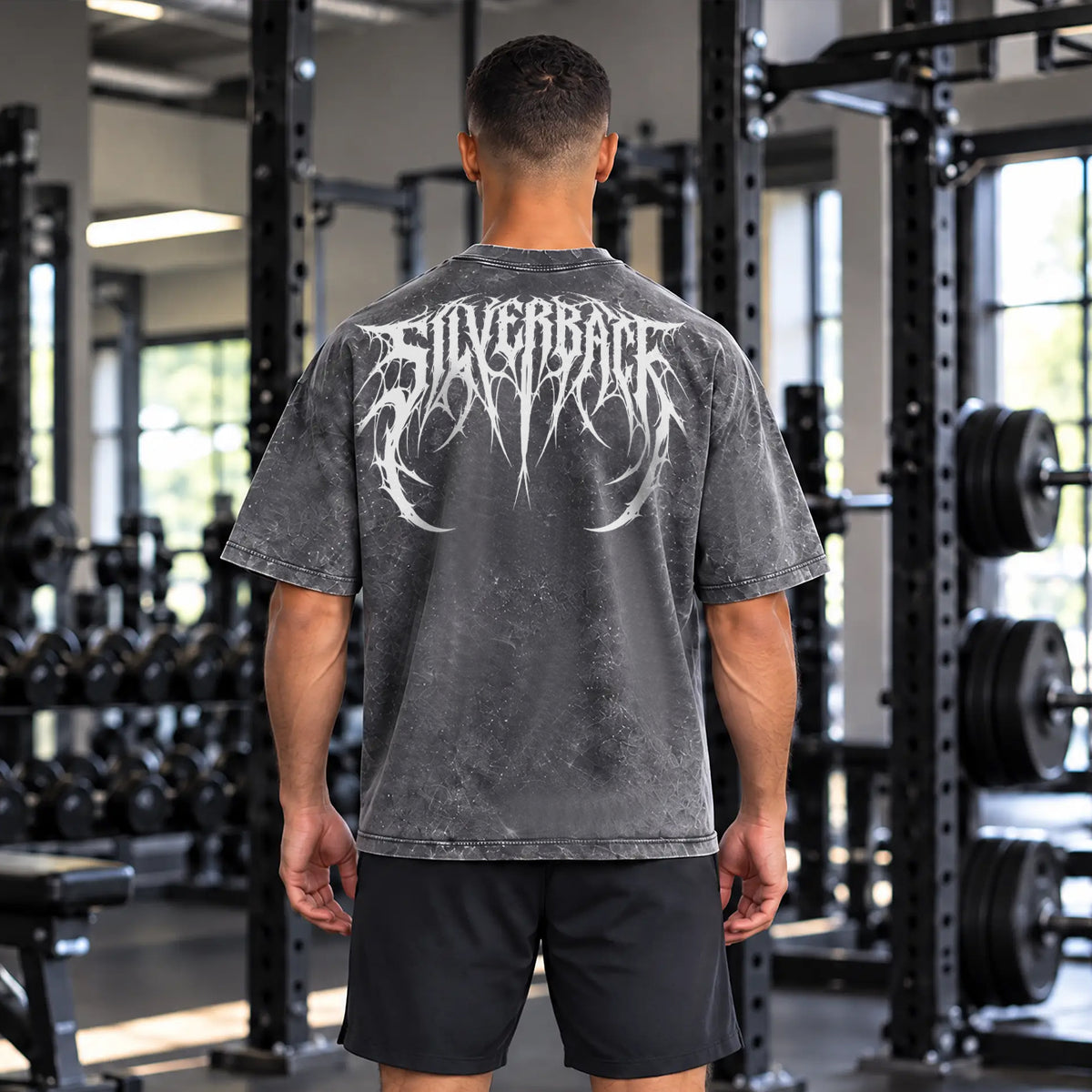A man viewed from behind in a gym, wearing a dark gray mineral-wash t-shirt with a large white 'SILVERBACK' band logo printed across the back, paired with black athletic shorts.